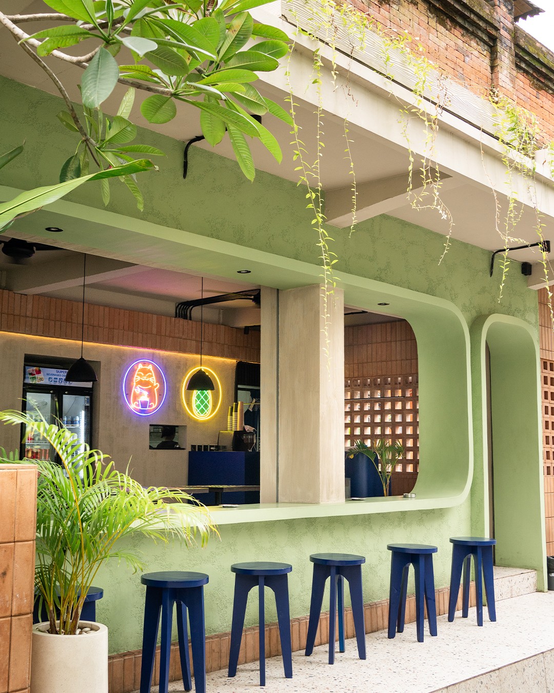 Bright green-themed cafe with neon signs, blue bar stools, hanging plants, and a wooden accented counter.